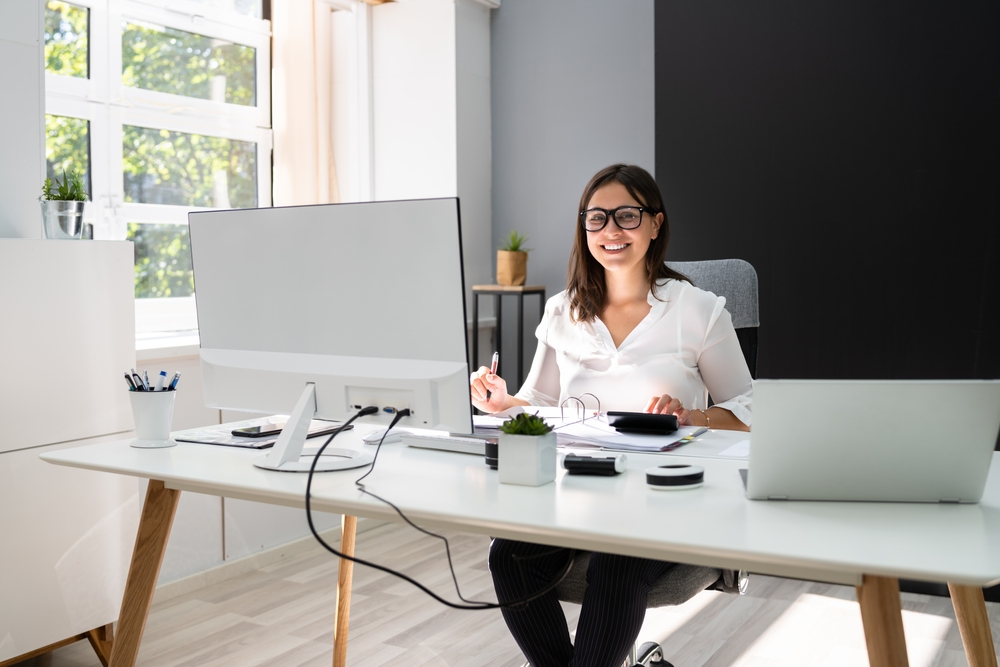 woman working in a nicely lit home office with huge monitor and a laptop